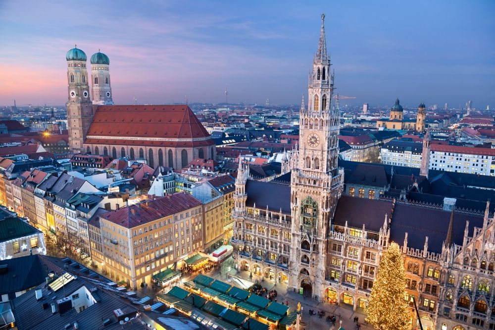 Auf dem Foto sieht man den Marienplatz von München aus der Vogelperspektive in der Dämmerung. Links im Bild ist die Frauenkirche zu sehen und weiter rechts im Bild sieht man das Rathaus. Unten im Bild sieht man einen beleuchteten Weihnachtsbaum und einige beleuchtete Marktstände. Start volunteering in Munich today!