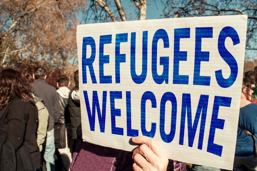 In the picture you can see somebody at a demonstration holding up a poster at a demonstration. On the poster it is written down "REFUGEE WELCOME" in blue letters. The people in the picture want to support and maybe are even going to be volunteering with refugees.