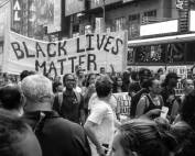 Auf dem Foto sind protestierende Menschen auf der Straße zu sehen. Das Foto ist in schwarz weiß. Die Menschen wollen sich gegen Rassismus engagieren. Auf einem großen Plakat steht in schwarzer Schrift "BLACK LIVES MATTER".