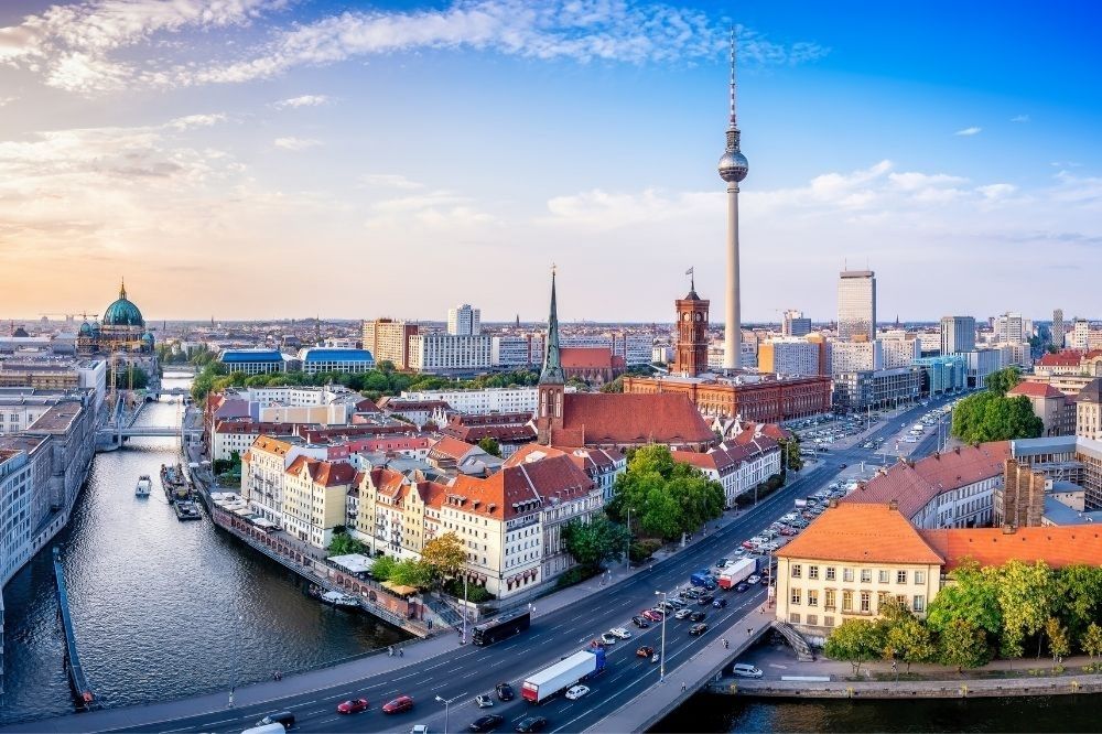 Auf dem Bild sieht man ein Panorama-Foto von Berlin. Links im Bild sieht man die Spree, über die eine Brücken mit Autos führt. Auf der rechten Seite sieht man den Fernsehturm. Das Foto entstand tagsüber und der Himmel ist blau und klar.