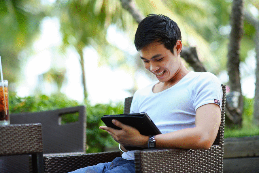 Auf dem Foto ist ein junger Mann in einem weißen T-Shirt zu sehen. Er sitzt draußen im Grünen und hält ein Tablet in der Hand.