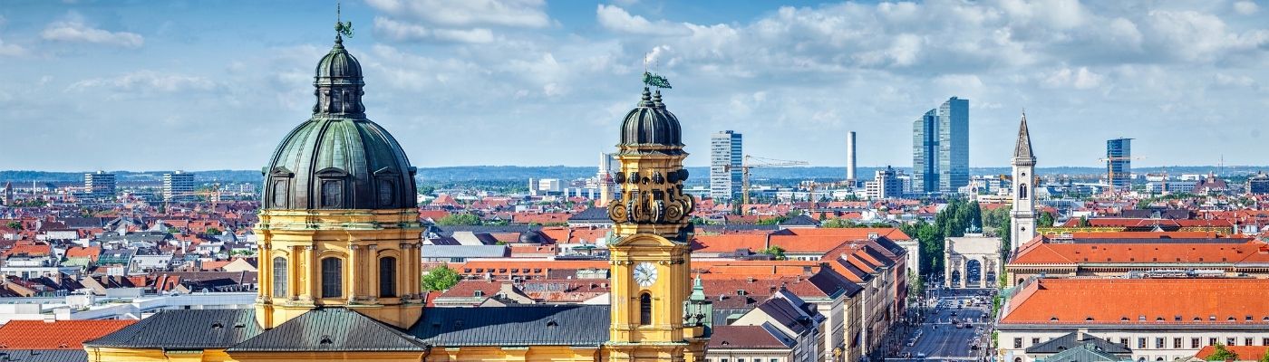 Auf dem Bild ist München zu sehen. Rechts im Bild ist die Frauenkirche mit ihren zwei Kirchtürmen mit grüner Kuppel zu sehen. Im Hintergrund sind Berge zu erkennen.