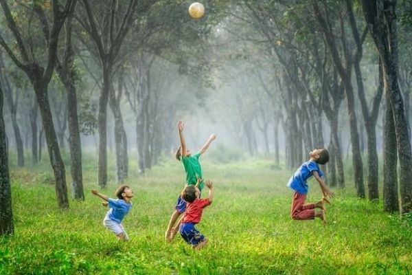 Auf dem Bild sieht man vier tobende Kinder auf einer Wiese, rechts und links sind Bäume. Sie springen in die Luft.