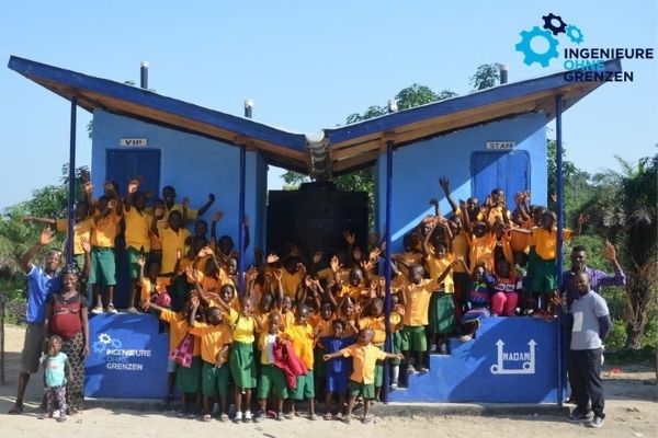 Auf dem Bild sieht man eine Gruppe von schwarzen Kindern in Sierra Leone vor einer erbauten Toilette. Rechts und links stehen erwachsene Menschen. Oben rechts ist das Logo der Organisation Ingenieure ohne Grenzen zu sehen.