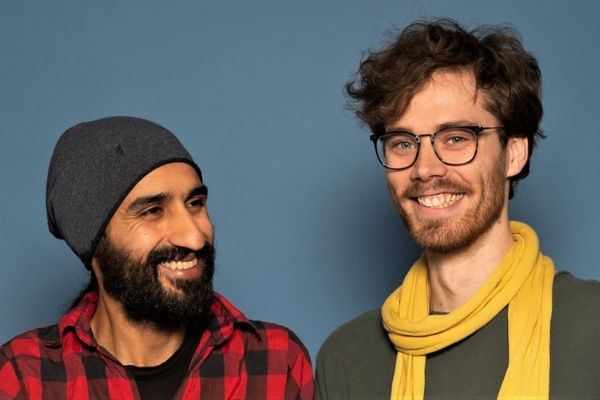 Auf dem Foto sieht man zwei junge Männer unterschiedlicher Herkunft vor einem blauen Hintergrund nebeneinander lachen.
