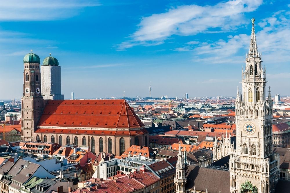 Das Bild zeigt München aus der Vogelperspektive. Man erkennt Gebäudedächer, der Himmel ist strahlend blau mit einigen Wolken.