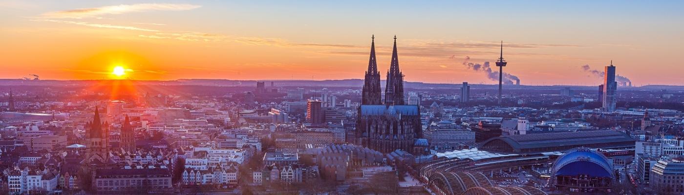 Auf dem Bild sieht man Köln als Panoramafotografie. Die Sonne geht gerade unter und von weitem erkennt man den kölner Dom in der Dämmerung.