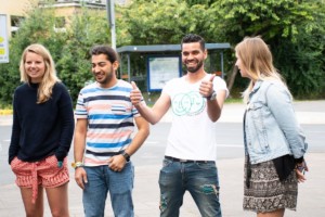 Zwei Männer und zwei Frauen sind draußen, umgeben von viel Grün. Sie lachen und sind glücklich. Einer hält seine beiden Daumen ausgestreckt und hat ein breites Lächeln.