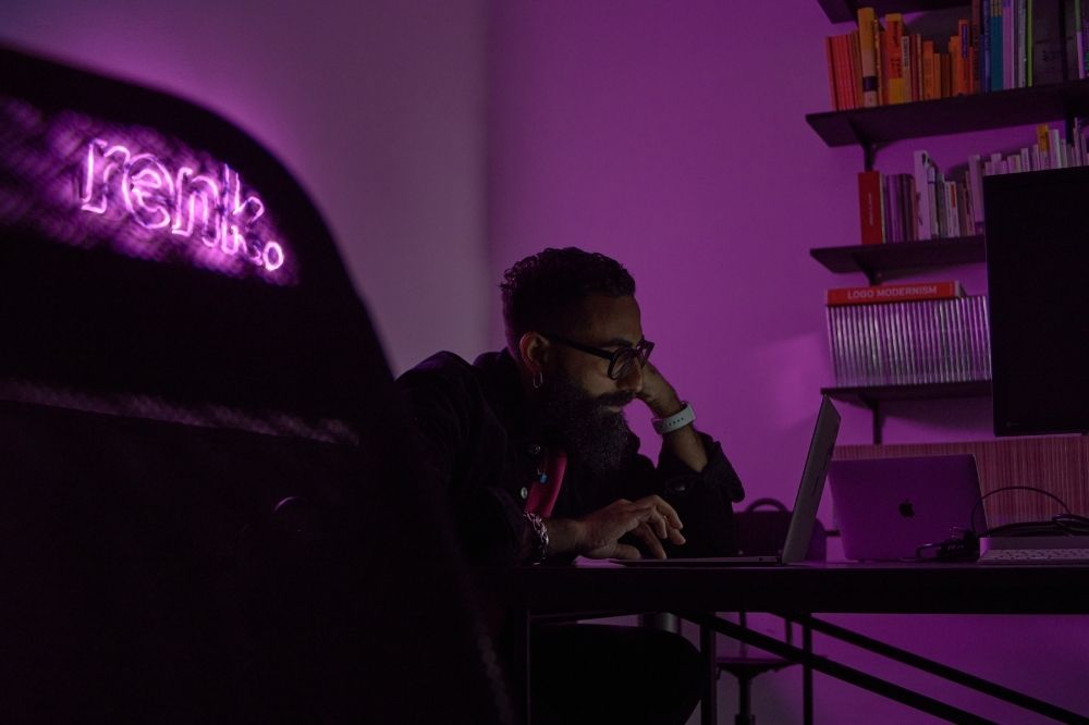 Auf dem Bild ist ein Mann vor einem Laptop zu sehen. Sein Name ist Erdal. Der Raum, in dem Erdal sitzt ist dunkel. Links im Bild steht in Leuchtschrift "renk". Die Schrift strahlt ein lilanes Licht aus, das den Raum erfüllt. Erdal setzt sich beim renk.magazin gegen Rassismus ein.