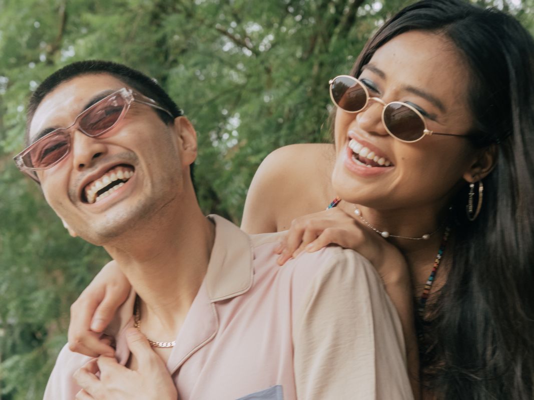 Ein Mann und eine Frau lachen. Beide haben Sonnenbrillen auf und sommerliche Kleidung an.