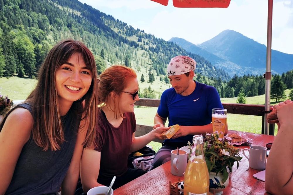 Auf dem Bild sieht man drei Personen, zwei Frauen und einen Mann, die an einem Tisch im freien sitzen und gemeinsam speisen. Der Tisch ist gedeckt und im Hintergrund sind grün und die Bergen zu sehen. Die Frau ganz vorne schaut und lacht in die Kamera.