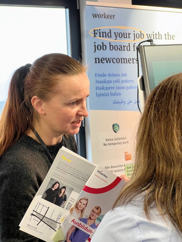 Olena bei ihrem Ehrenamt. Sie spricht an einem Stand bei einer Jobbörse mit einer anderen Person und hat einen Flyer in der Hand.