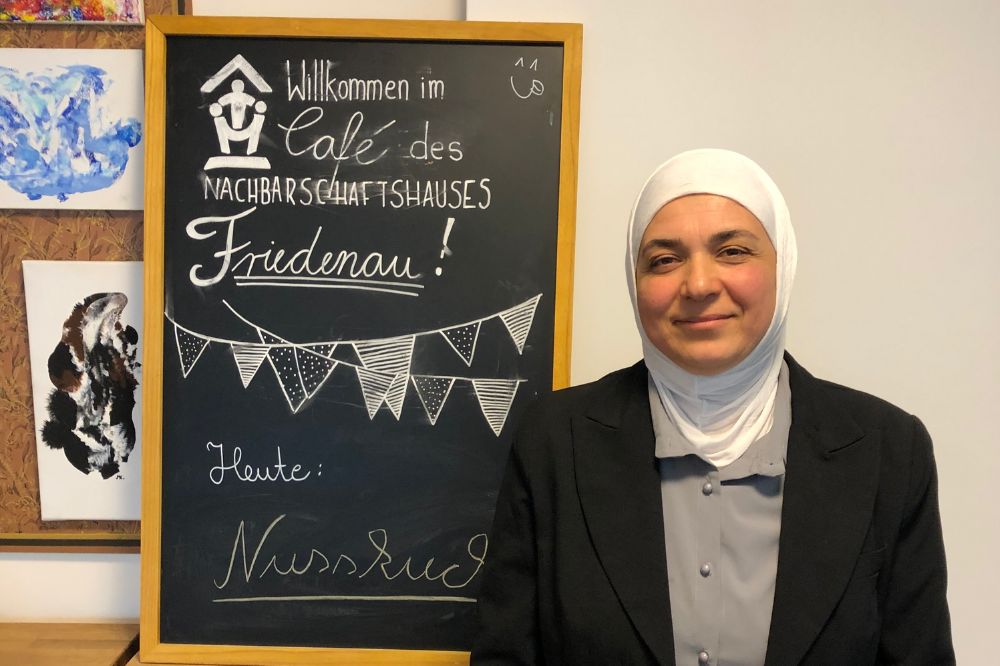 Auf dem Bild ist Khalsa im rechten Bildrand zu sehen. Sie befindet sich in einem Café. Links neben Ihr ist eine Tafel mit der Aufschrift: "Willkommen im Café des Nachbarschaftshauses Friedenau. Heute: Nusskuchen". Khalsa lächelt in die Kamera.