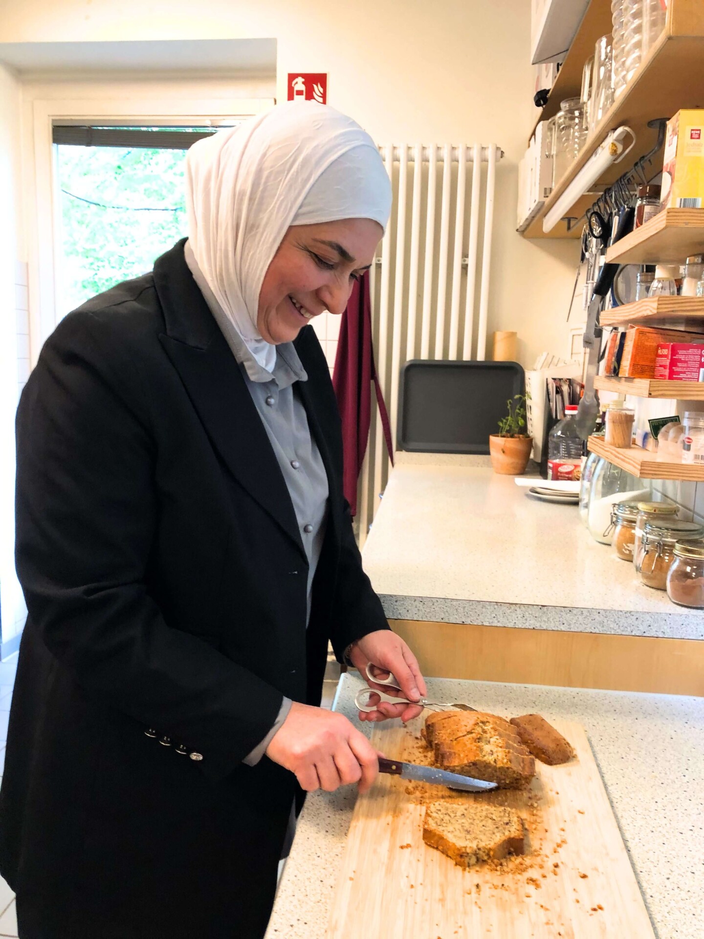 Khalsa befindet sich in der Küche und schneidet gerade auf einem Schneidebrett einen Kuchen in Scheiben. Sie lächelt.