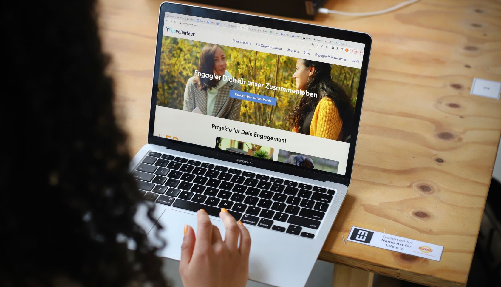 Aufgeklappter Laptop, davor sitzt eine Person mit dem Rücken zur Kamera. Auf dem Bildschirm sieht man die GoVolunteer-Website.