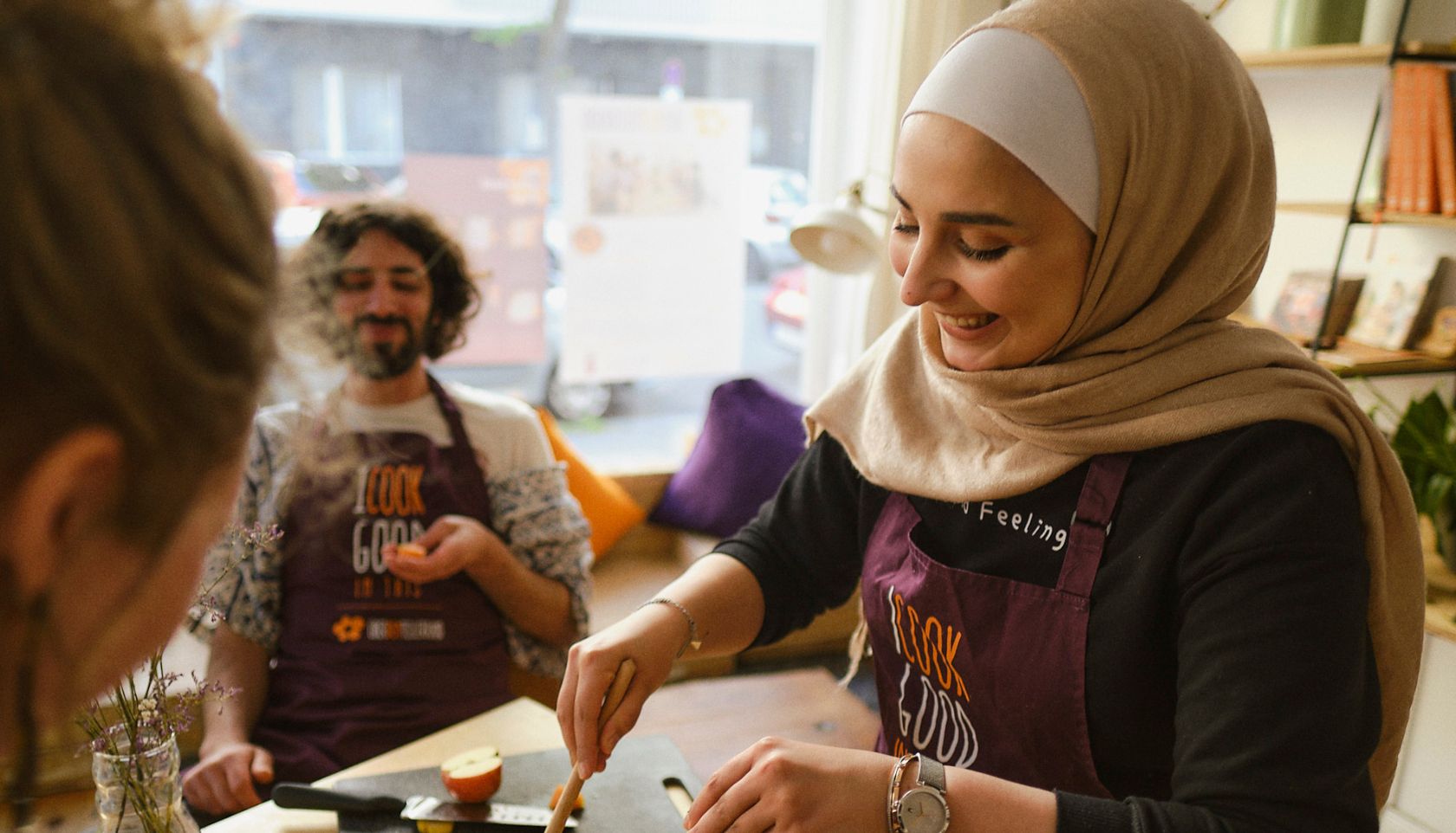 Menschen kochen gemeinsam, im Vordergrund eine Frau mit Kopftuch, die lächelt. Im Hintergrund lacht ein Man..